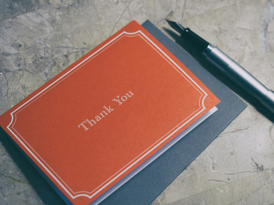Orange 'Thank You' card on a textured surface with a pen.